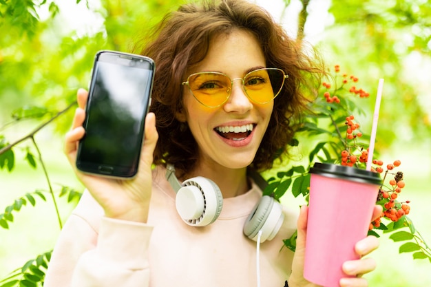 Photo pretty young caucasian woman go for a walk in the park with cup of coffee and show her phone