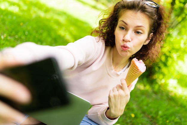 Pretty young caucasian woman is engaged in freelance in the park with laptop, the cup of coffe and ice-cream, sits on a grass and does selfie