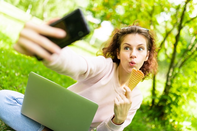 Pretty young caucasian woman is engaged in freelance in the park with laptop, the cup of coffe and ice-cream, sits on a grass and does selfie