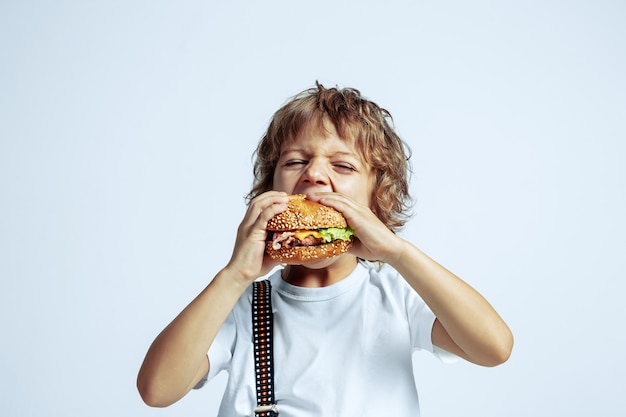 Pretty young curly boy in casual clothes on white wall. Eating burger. Caucasian male preschooler with bright facial emotions. Childhood, expression, having fun, fast food. Hungry.