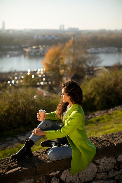 Pretty young curly hair woman enjoying autumn sun while sitting by the river and drinking takeaway coffee