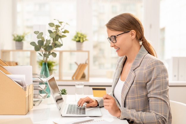 Pretty young elegant businesswoman with plastic card sitting in front of laptop while searching for goods in online shop