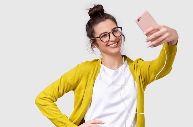 Pretty young European woman in casual clothes standing and taking a selfie isolated over white studio background Pretty Caucasian brunette female wearing transparent spectacles making self portrait