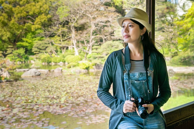 pretty young female visitor holding a camera and looking outside while sitting near the window in a garden house.