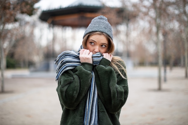 Photo pretty young female in warm clothes wrapping in scarf and looking away in autumn park on really cold day