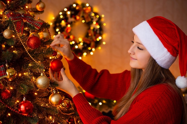 Pretty young girl decorating Christmas tree before winter holidays Winter time family at home happy atmosphere warm bright garland light in the room