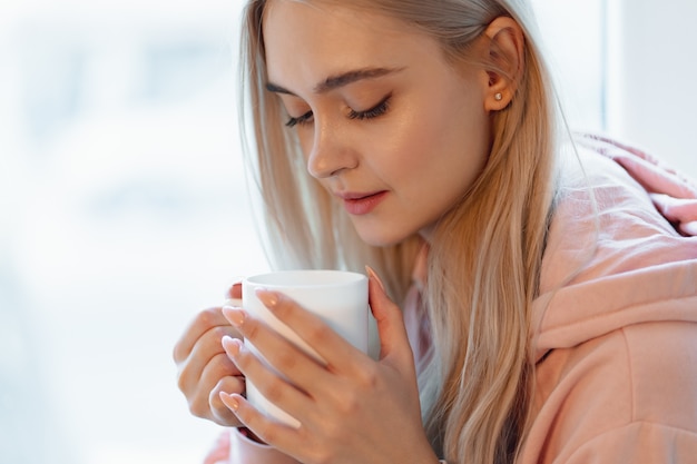 A pretty young girl enjoying a cup of coffee or milk while sitting by the window. Wearing a casual pink, pale parka. 