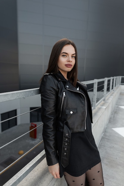 Pretty young girl in fashionable black clothes with a leather jacket and dress stands near a metal fence in the city