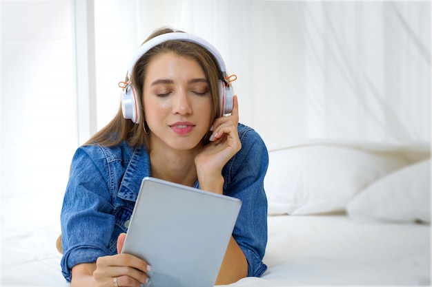 Pretty young girl is listening the music from tablet on the white bed