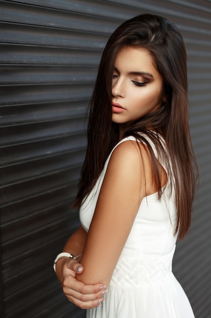 Pretty young girl with makeup in white dress near metal dark wall