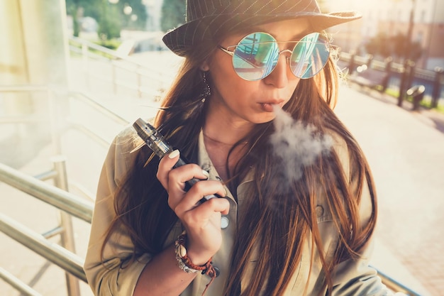 Pretty young hipster woman in black hat vape ecig vaping device at the sunset Toned image