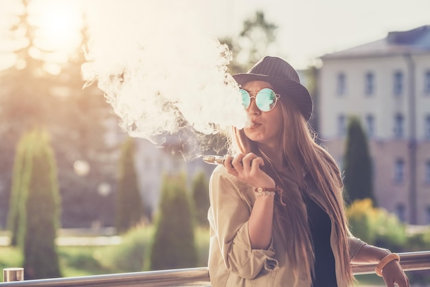 Pretty young hipster woman in black hat vape ecig vaping device at the sunset Toned image