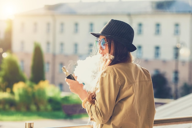 Pretty young hipster woman in black hat vape ecig vaping device at the sunset Toned image