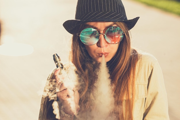 Pretty young hipster woman in black hat vape ecig vaping device at the sunset Toned image