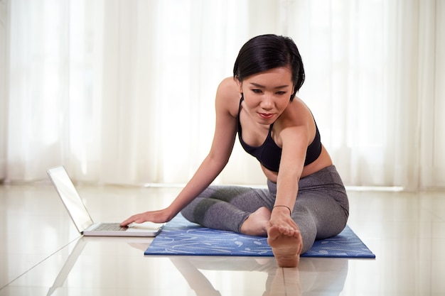 Pretty young Indian woman following tutorial on laptop screen when doing stretching exercise