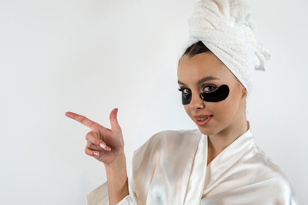 Pretty young lady in white towel applying collagen hydrogel patches under the eyes to smooth the skin isolated on white