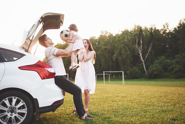 Pretty young married couple and their daughter are resting in the nature. The mother father and little girl are sitting on open car boot