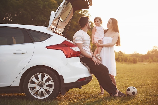 Pretty young married couple and their daughter are resting in the nature. The mother father and little girl are sitting on open car boot