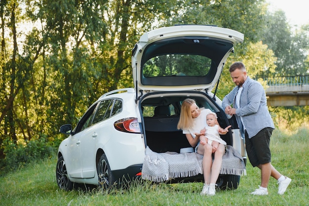 Pretty young married couple and their daughter are resting in the nature. The woman and girl are sitting on open car boot. The man is standing near them. They are smiling