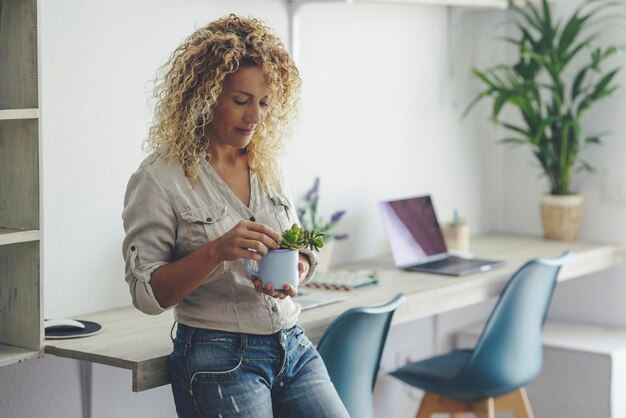 Pretty young mature lady woman enjoy indoor leisure activity at home standing and caring a plant Laptop computer in background as alternative office workstation Modern cute lady in casual wear smile