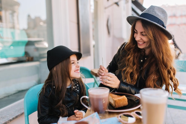 Pretty young mother and her cute daughter delicious breakfast with coffee and dessert in a cafe on the city background. Styling family, true emotions, good day.