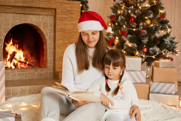 Pretty young mother reading book to her daughter while sitting near fireplace and fir tree in festive living room