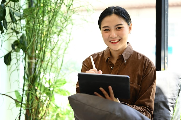 Pretty young office worker sitting on armchair in modern office and using digital tablet