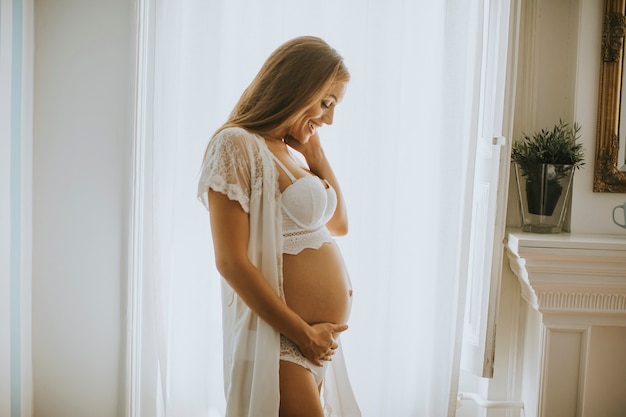 Pretty young pregnant woman standing at window in the room at daylight