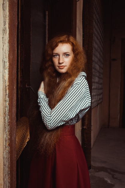 Pretty young red haired model wearing white striped shirt and red skirt. Woman posing in a dark room