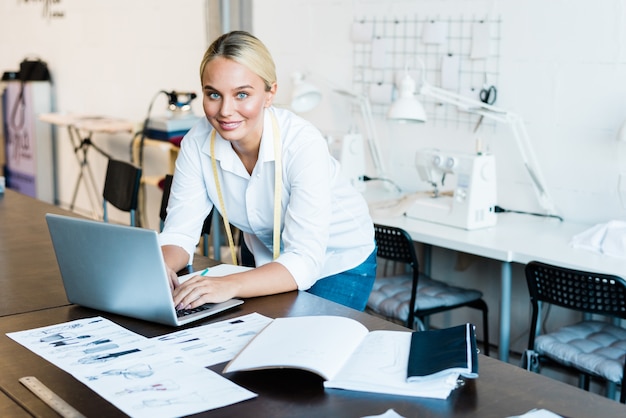 Pretty young smiling fashion designer in casualwear looking at you while bending over laptop and scrolling through online stuff