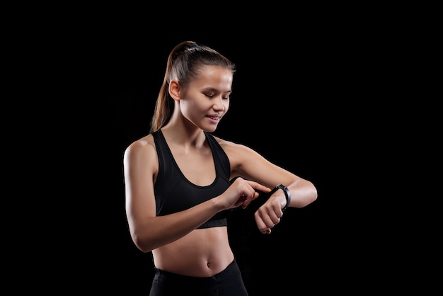 Pretty young smiling sportswoman in activewear looking at her smartwatch and pointing at its display while standing in isolation