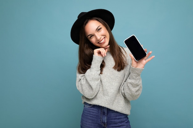 Pretty young smiling woman wearing black hat and grey sweater holding phone