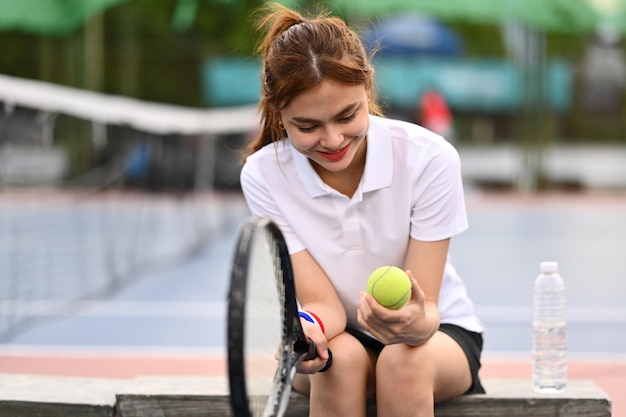 Pretty young sportswoman holding ball racket sitting on the bench at tennis court