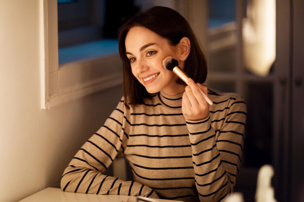 Photo pretty young woman applying blush with brush home interior