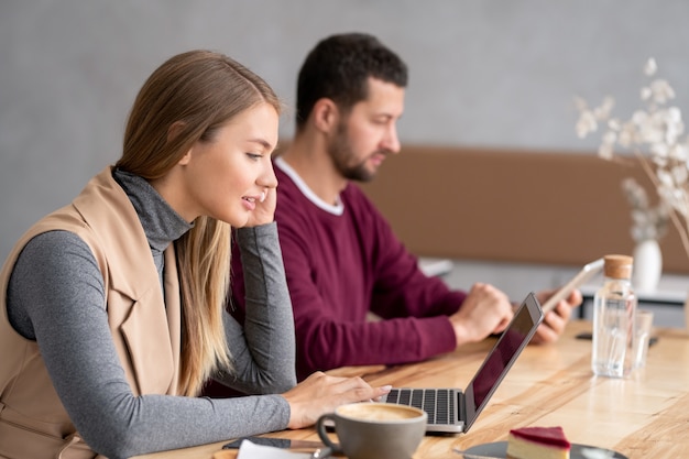 Pretty young woman in casualwear sitting by wooden table in front of laptop display and communicating through video-chat