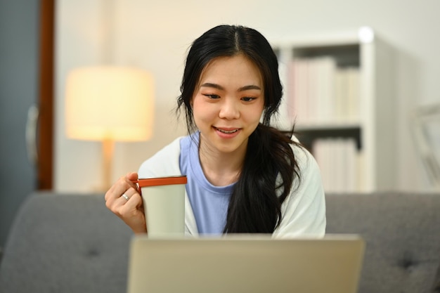 Pretty young woman drinking herbal tea on couch and watching video or reading email on laptop