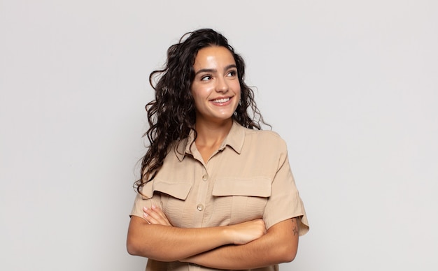 Pretty young woman feeling happy, proud and hopeful, wondering or thinking, looking up to copy space with crossed arms