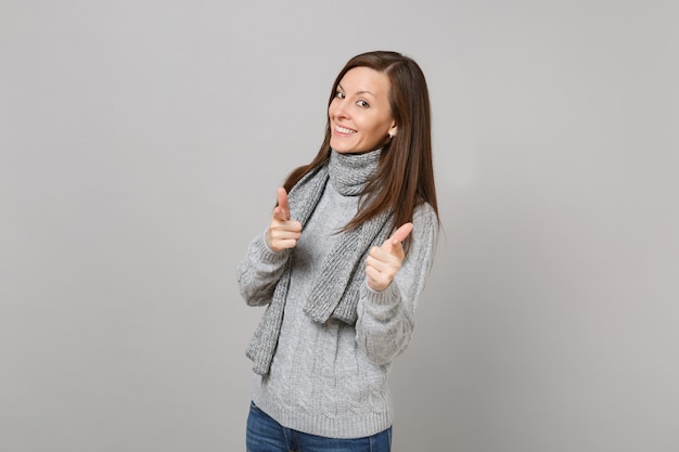 Pretty young woman in gray sweater, scarf pointing index fingers on camera isolated on grey wall background. Healthy fashion lifestyle, people sincere emotions cold season concept. Mock up copy space.