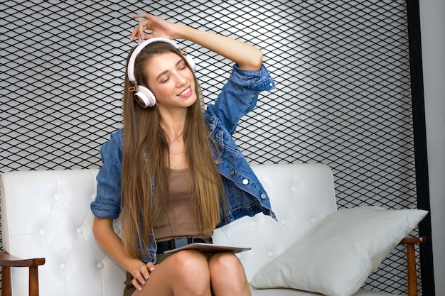 Pretty young woman is sitting on sofa and wearing headphone for listening the music from tablet