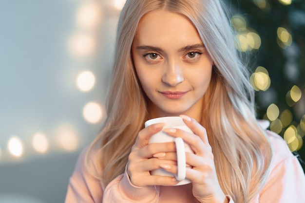 A pretty young woman making a wish while drinking a hot cup of milk, coffee waiting holidays with blurred lights on tree in the background at home.