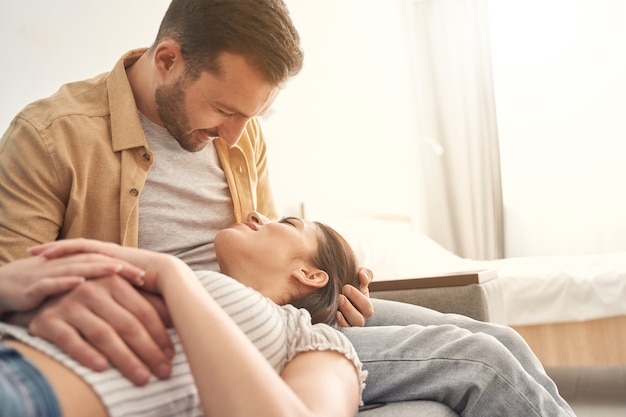 Pretty young woman resting on knees of her boyfriend while staying in living room at home