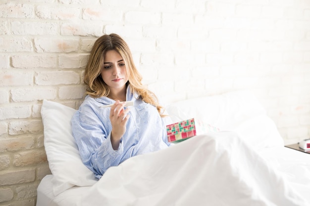 Pretty young woman in sitting in bed while reading a thermometer and holding a box of tissues