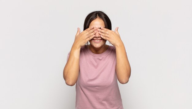 Pretty young woman smiling and feeling happy, covering eyes with both hands and waiting for unbelievable surprise