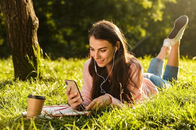 Pretty young woman student using mobile phone while laying on a grass at the park, listening to music with earphones