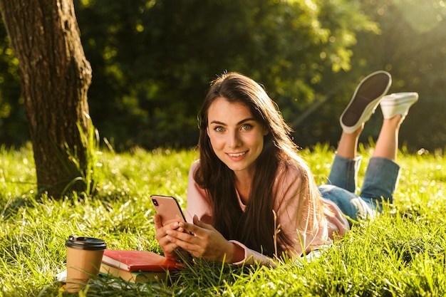 Pretty young woman student using mobile phone while laying on a grass at the park