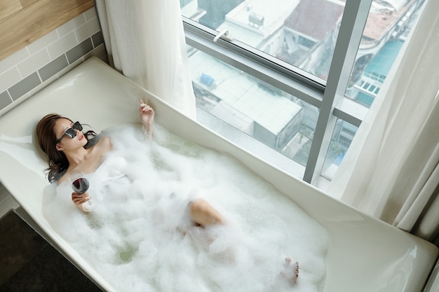 Pretty young woman in sunglasses relaxing in bubble bath with glass of wine, view from above