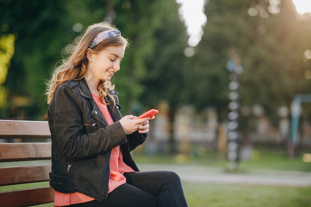 Pretty young woman in trendy casual outfit sits on a bench in the city park. Being online, modern technologies