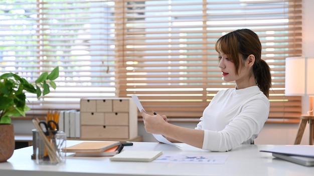 Pretty young woman in white sweater reading document and using laptop during working from home