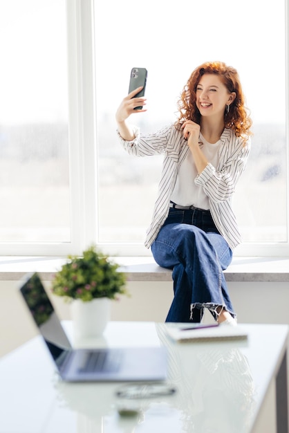 Pretty young woman with long red hair takes a selfie on her smartphone while sitting on the windowsill Beautiful girl with a phone in a home environment Technology and social networks concept