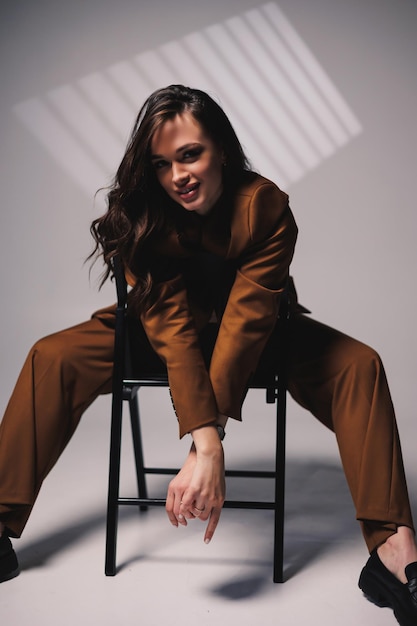 A pretty young woman with a smile on her face and wearing a brown classic suit is sitting on a chair on a gray background Beautiful woman in a jacket and with makeup on her face